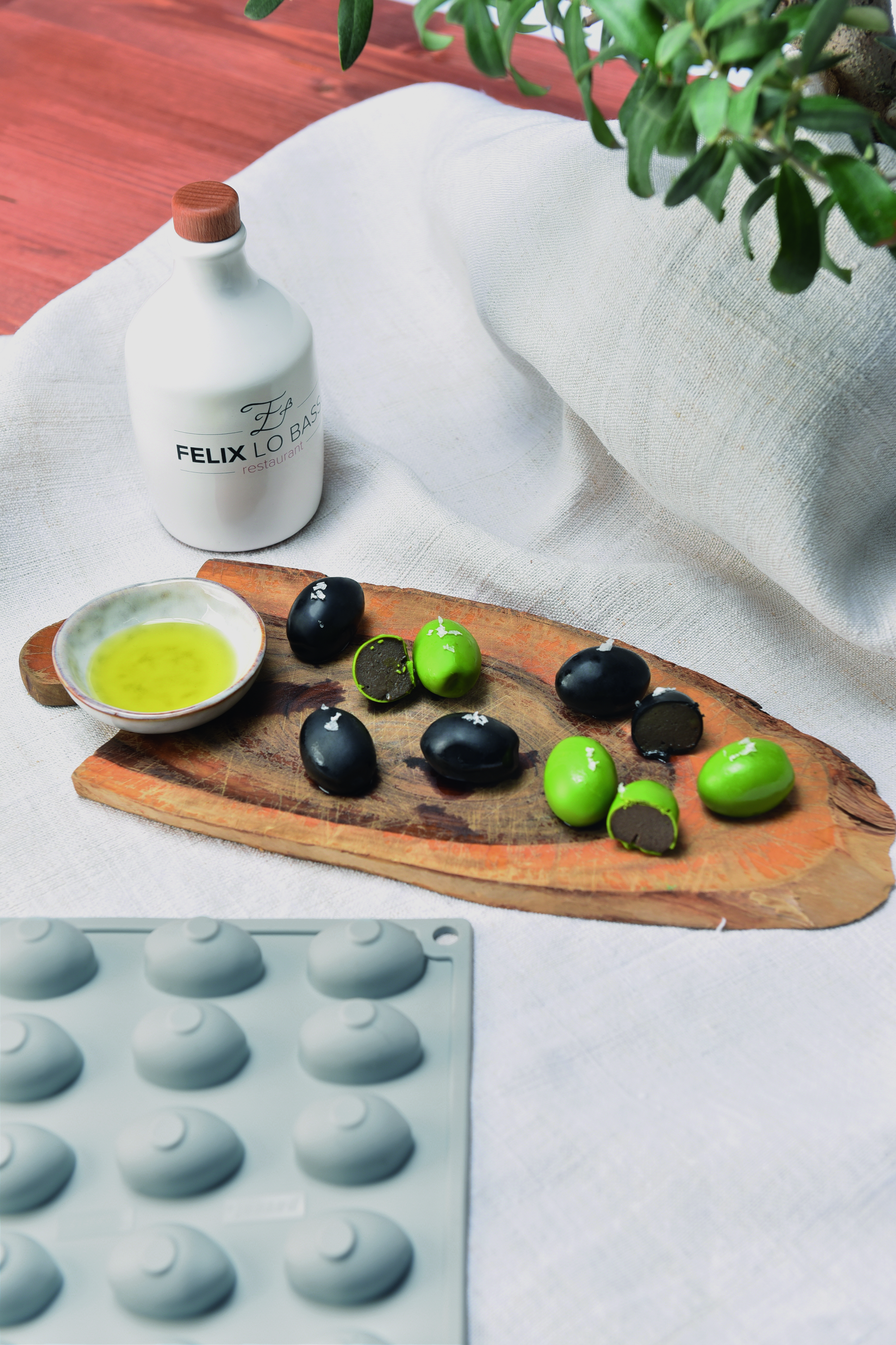 Holzbrettchen mit grünen und schwarzen Oliven und kleinem Schälchen mit Olivenöl auf weißer Tischdecke, Silikonform Oliven unten rechts davor
