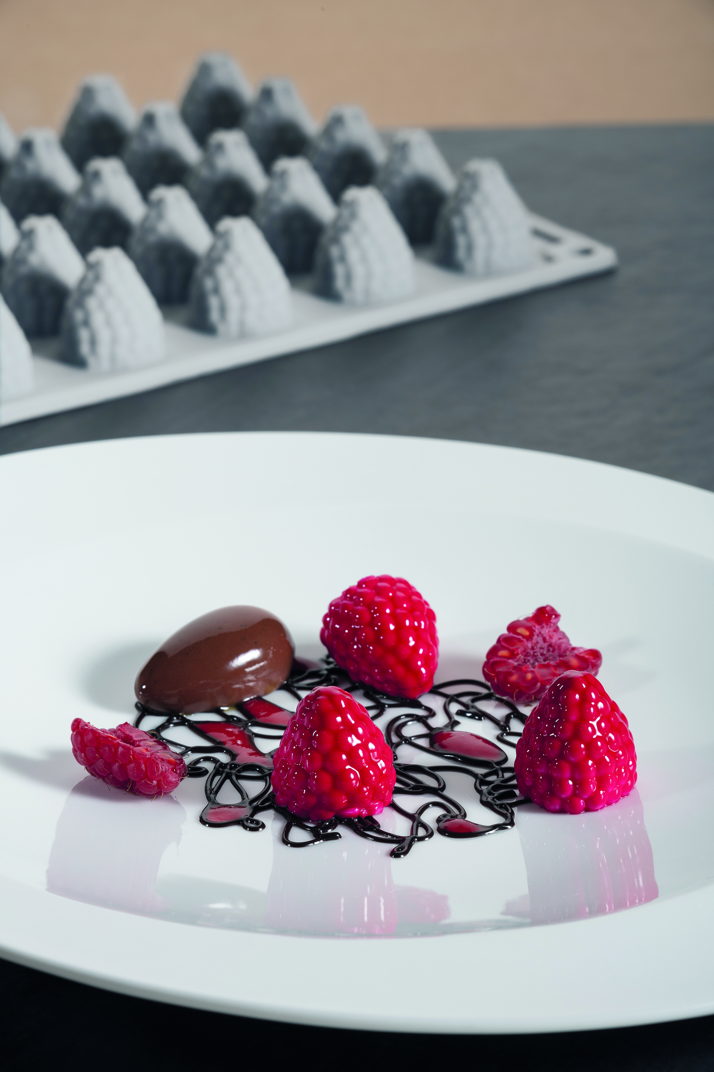 weißer Porzellanteller mit Himbeeren aus Silikonform Beeren, GG017S, mit Schokoladenkringeln und einem mandelförmigen Stück Schokolade, im Hintergrund Silikonform Beeren GG017S